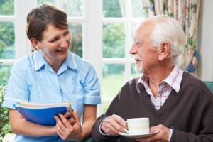 A caregiver and senior sitting together at a table with photos, talking and smiling in a sunlit common room.